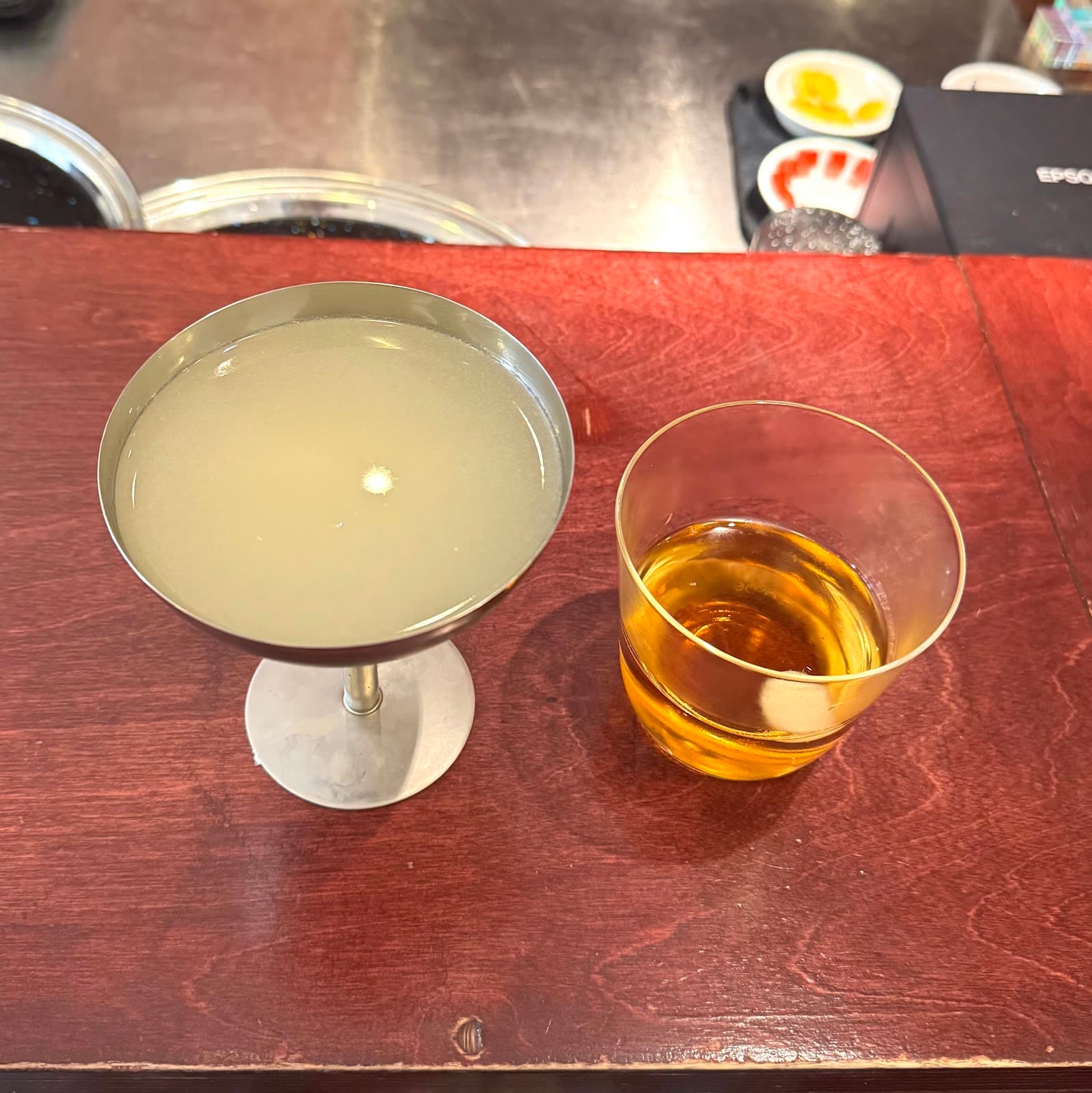 Two cocktails, one pale in a coupe glass and one amber in a rocks glass, on a wooden bar