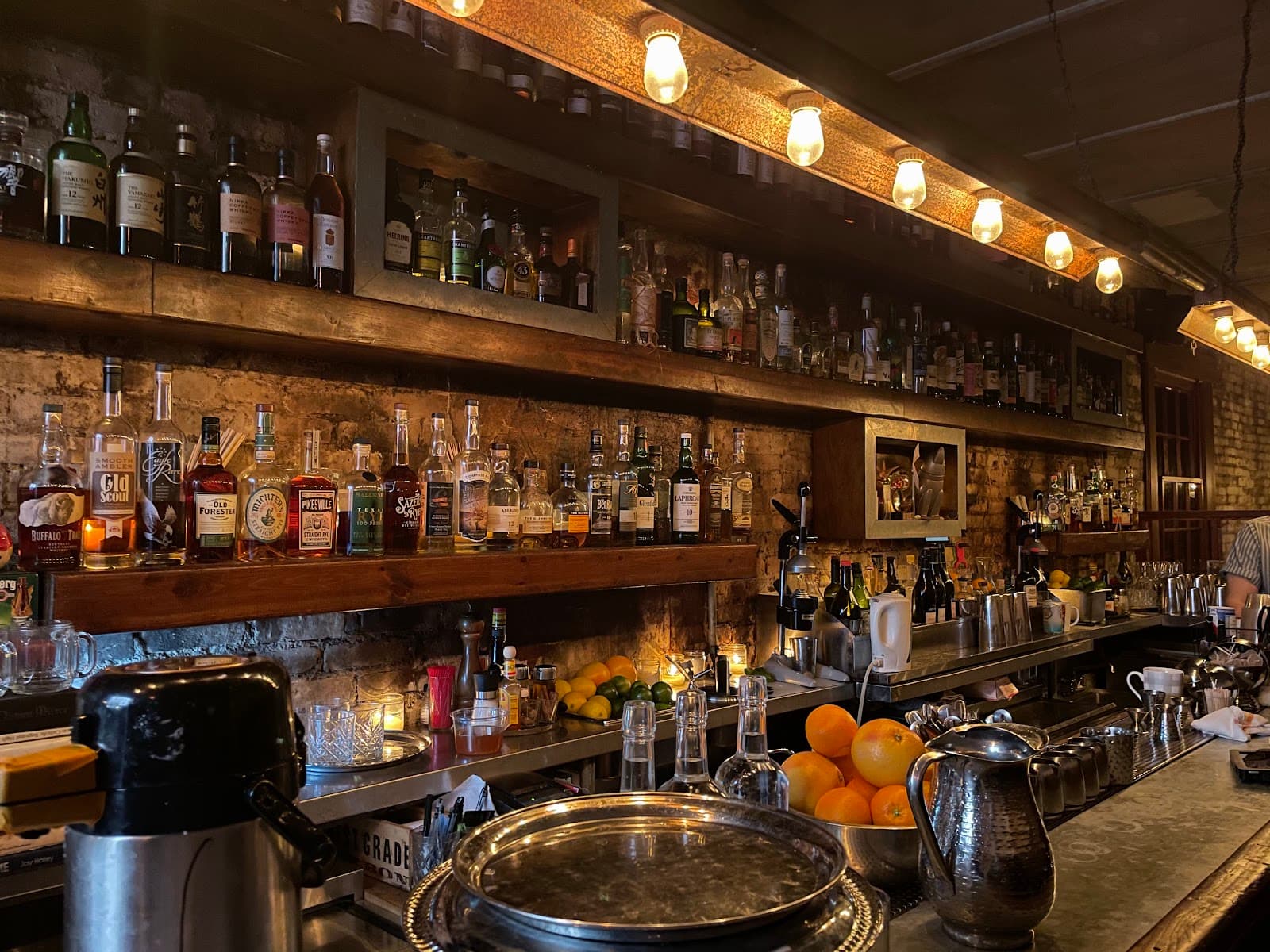 Warmly lit bar with shelves of liquor bottles and a countertop with oranges and equipment