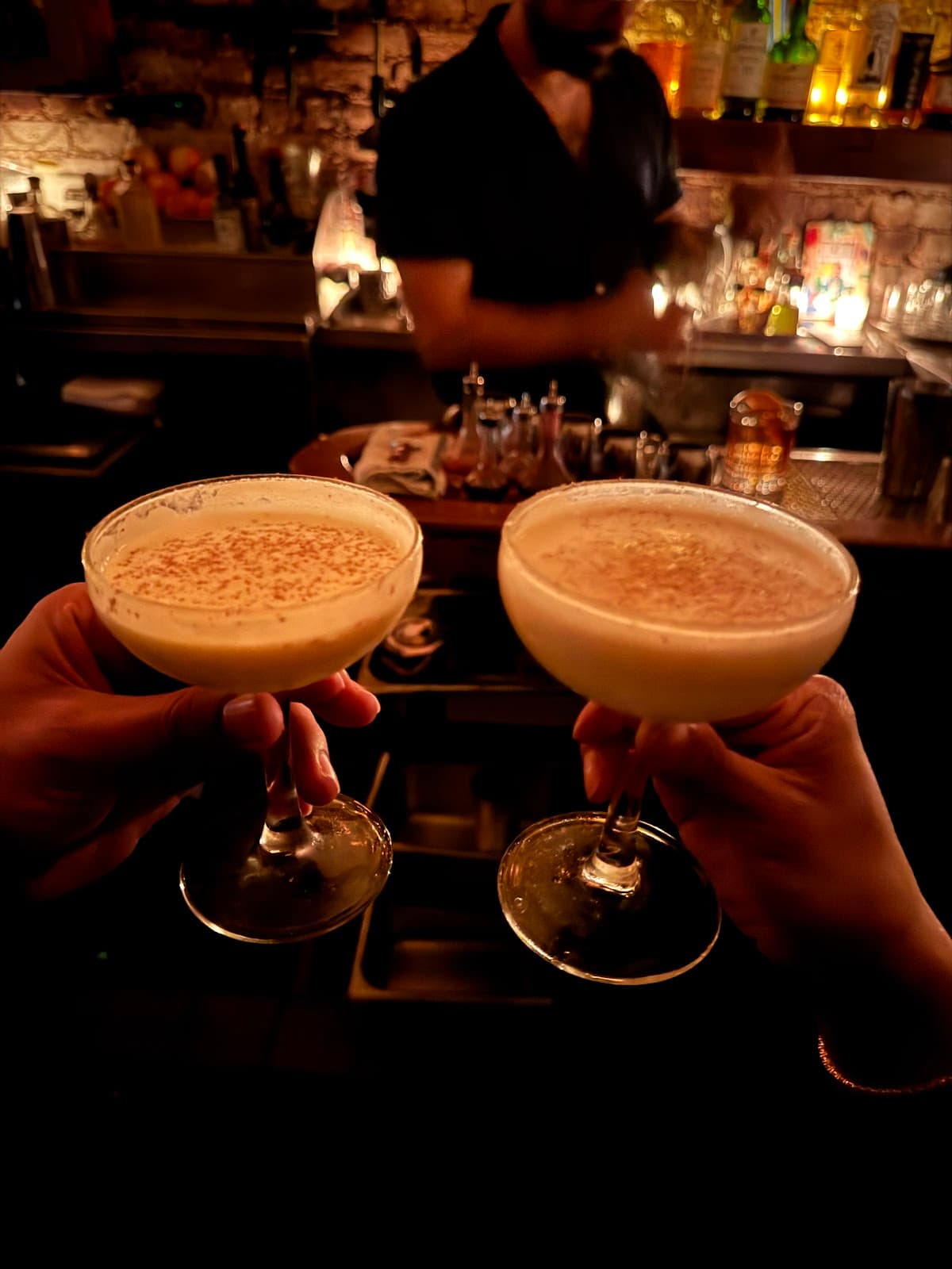 Two hands holding cocktails in a dimly lit bar atmosphere in New York