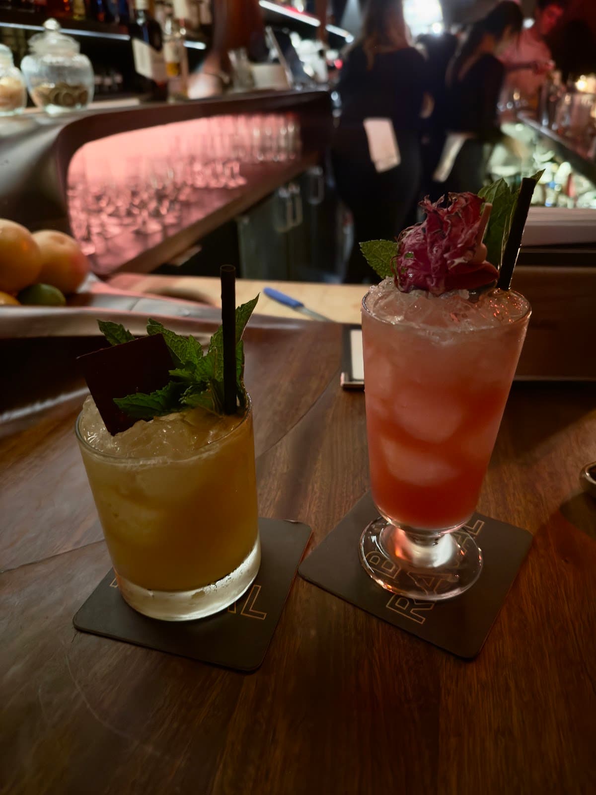 Two craft cocktails with garnishes on a bar counter in a dimly lit setting in Toronto
