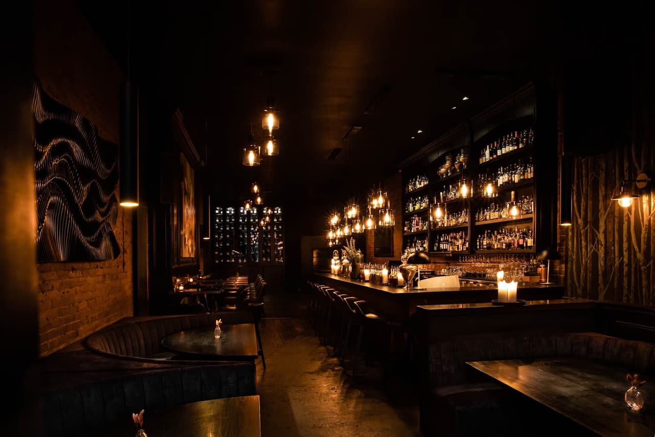 Warmly lit interior of a cocktail bar with wooden bar counter and shelves of bottles