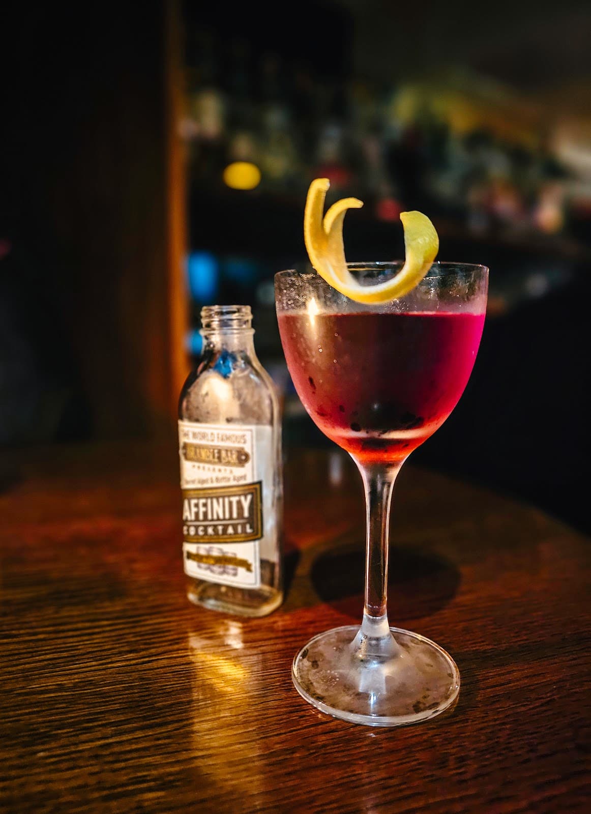 Cocktail glass with red drink and lemon twist beside bottle on a wooden bar table in Edinburgh