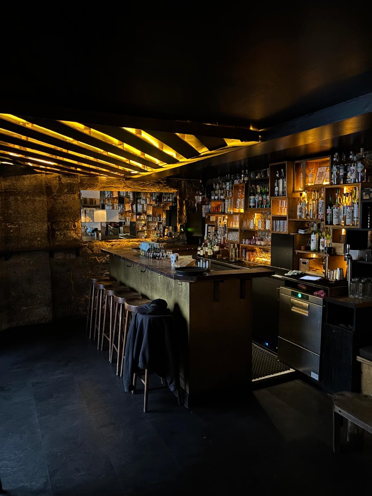 Dimly lit bar counter with shelves of bottles and wooden stools in Paris