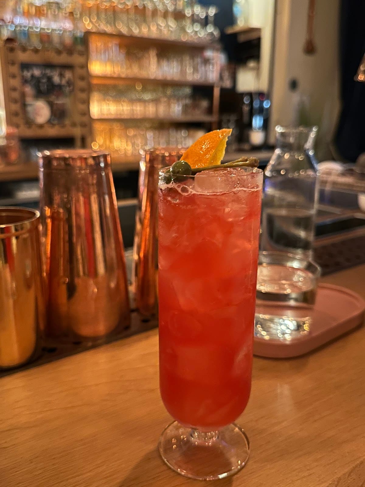 Orange and caper garnished cocktail on a bar counter with glassware in background in Paris