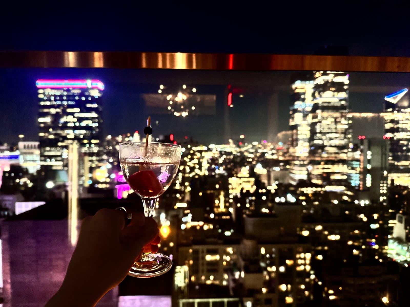 Cocktail glass held against a city skyline at night with colored lights in New York