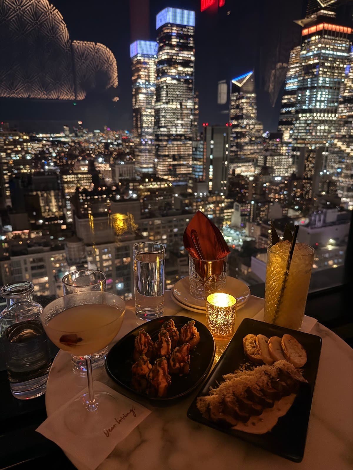 Nighttime city view with cocktails and appetizers at a table by the window. in New York