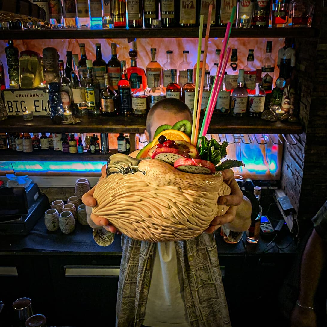 Bartender holding a large cocktail with fruit garnish in front of a bar shelf in Paris
