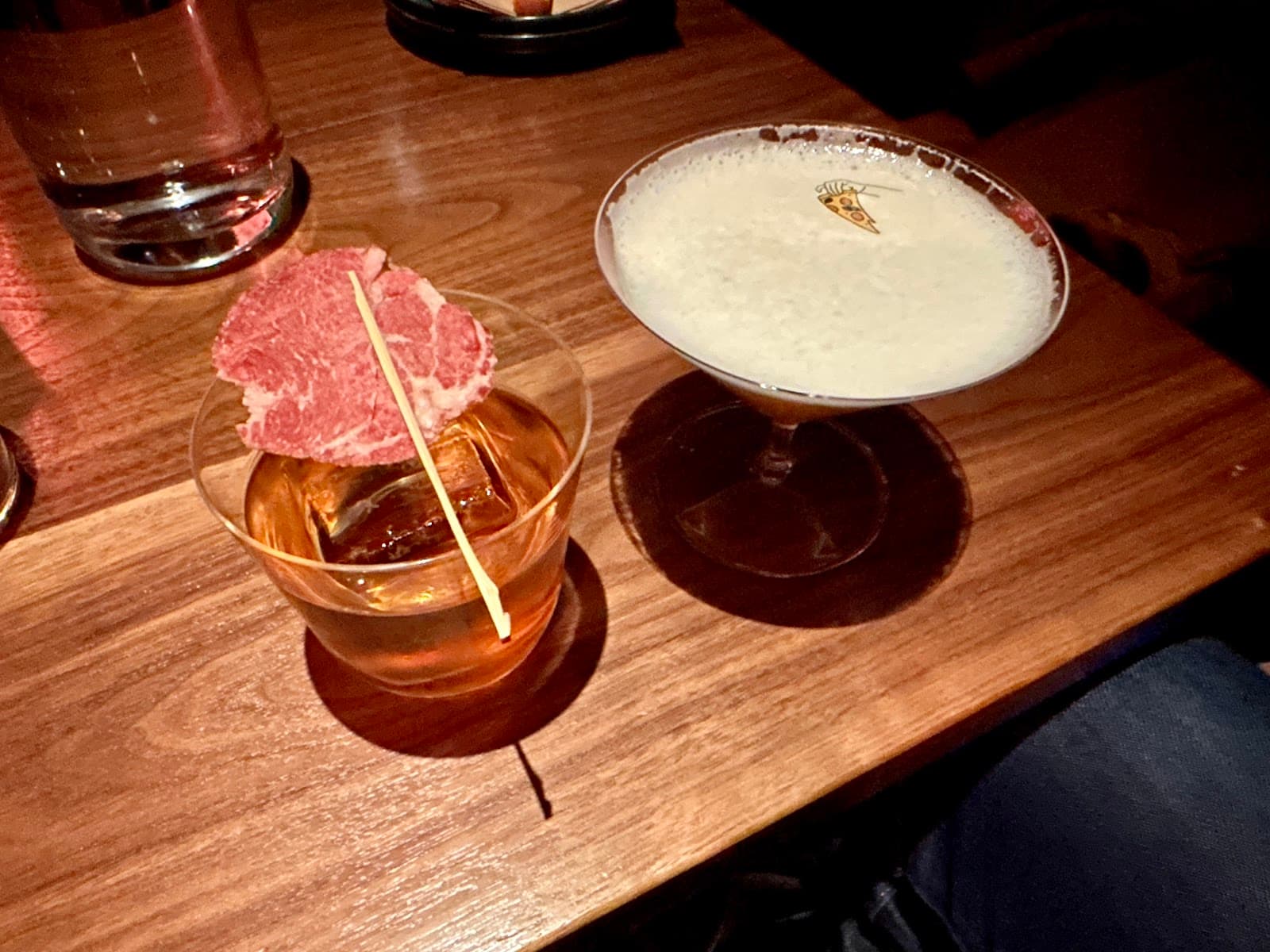 Two cocktails on a wooden table, one with a slice of meat garnish. in New York