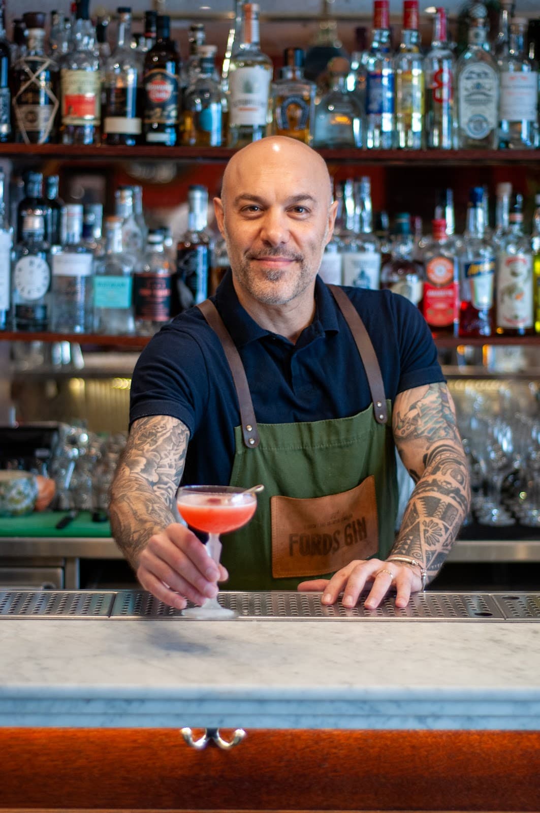 Bartender with a tattoo serving a cocktail at a bar with bottles in the background in London