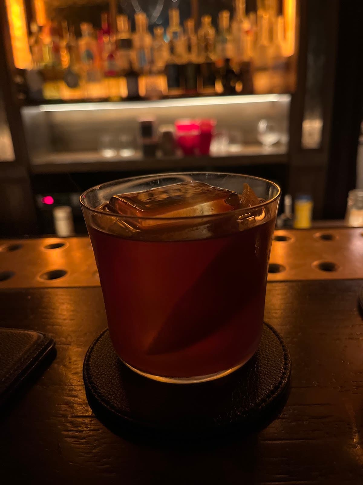 Dark cocktail on bar with ice cube and blurred bottles in the background in London
