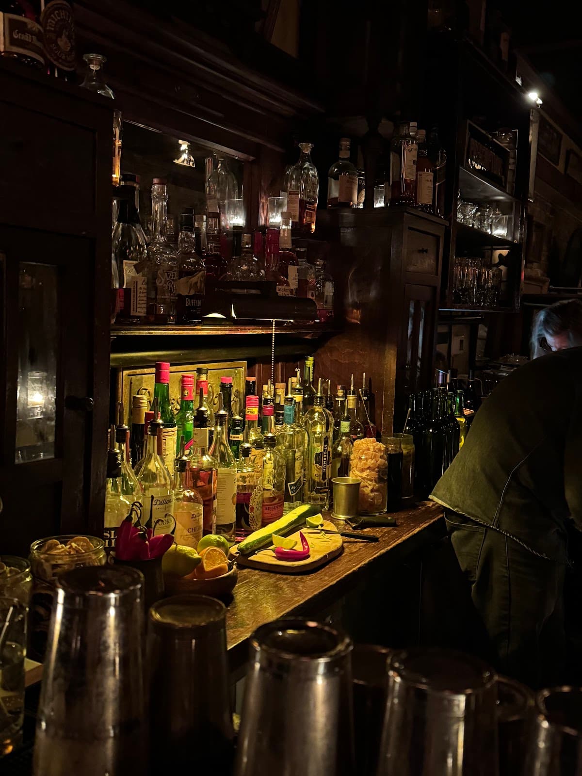 Dimly lit bar counter with bottles and cutting board with limes and cucumbers