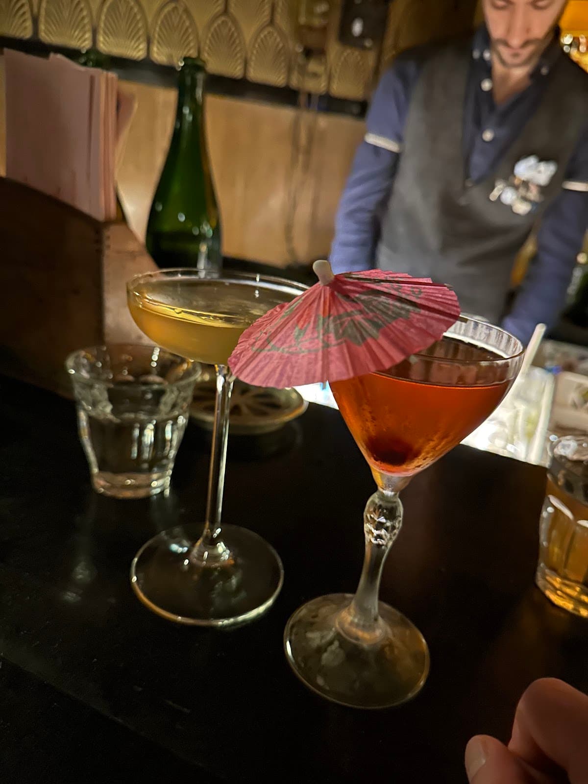 Close-up of two cocktails on a dimly lit bar counter with a bartender in the background in Paris