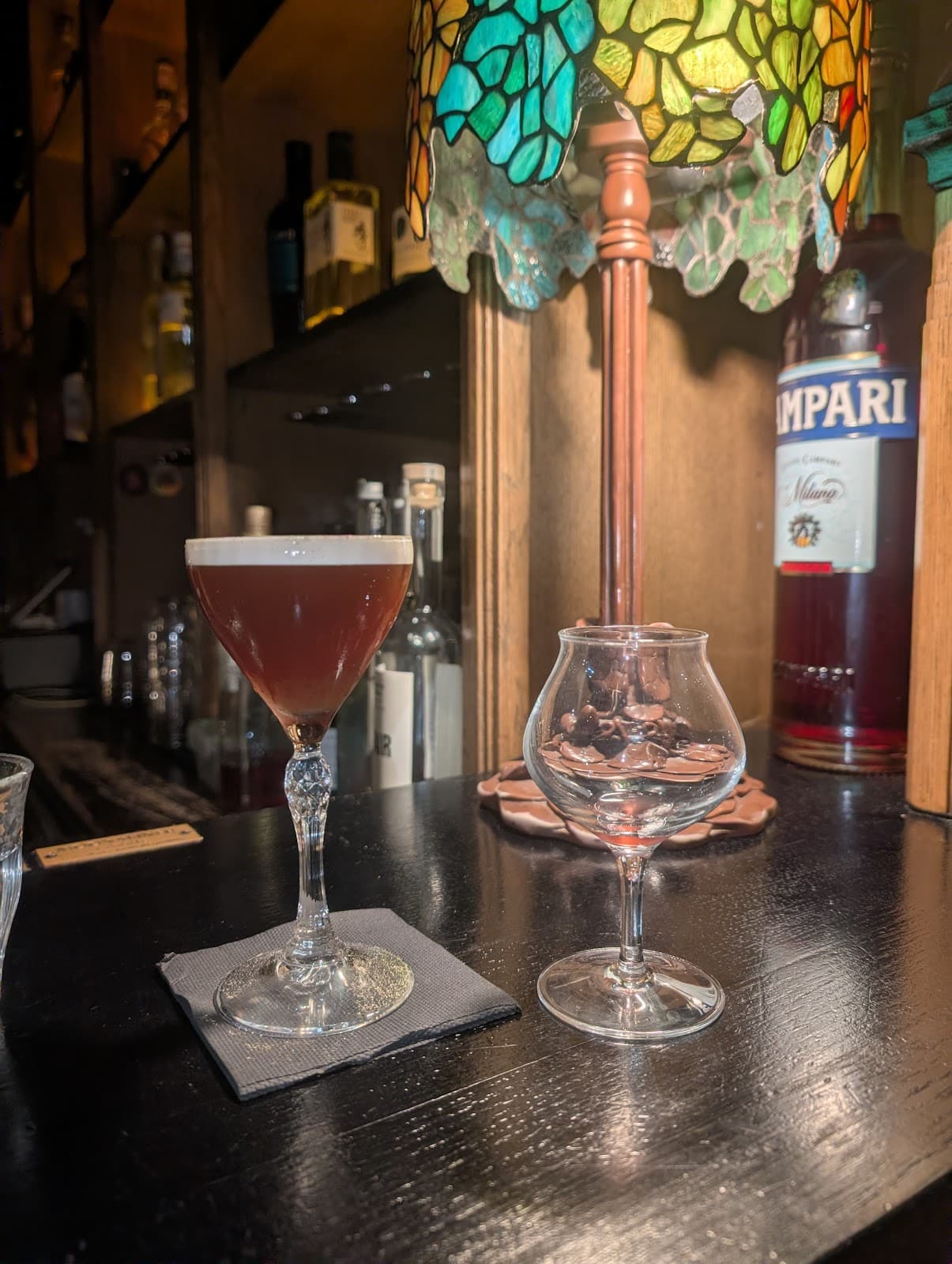 Cocktail glass on a bar with a colorful lamp and bottles in the background in Paris
