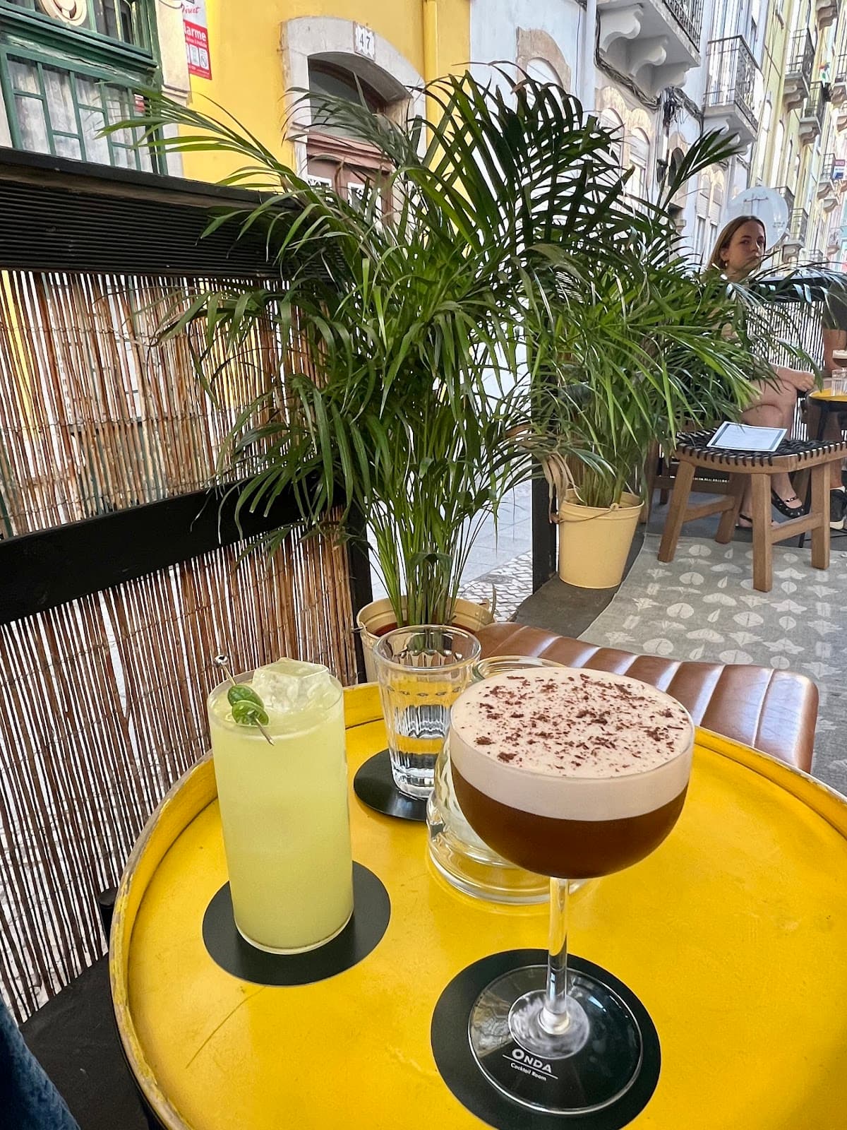 Two cocktails on a yellow table, with plants and street view background in Lisboa
