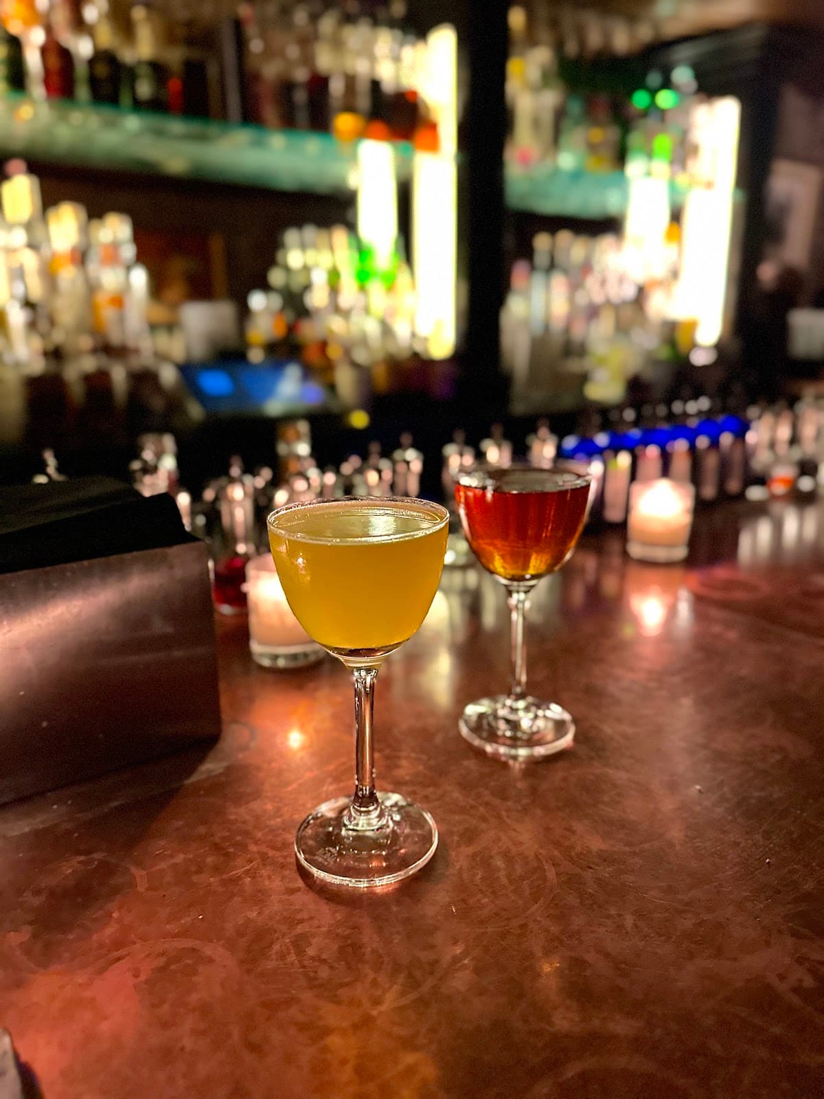 Two cocktails on a dimly lit bar counter with blurred bottles in background in New York