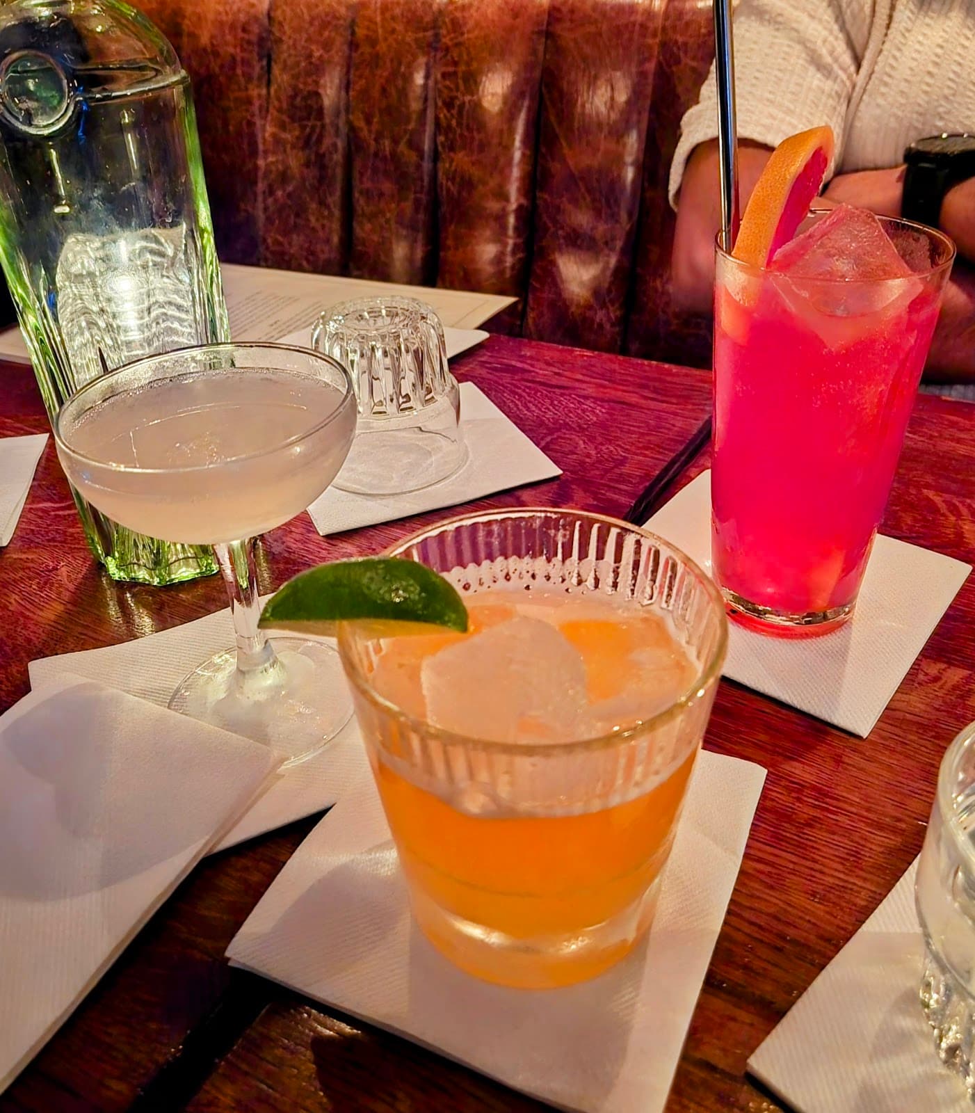 Three cocktails on a bar table with napkins and a glass bottle. in London