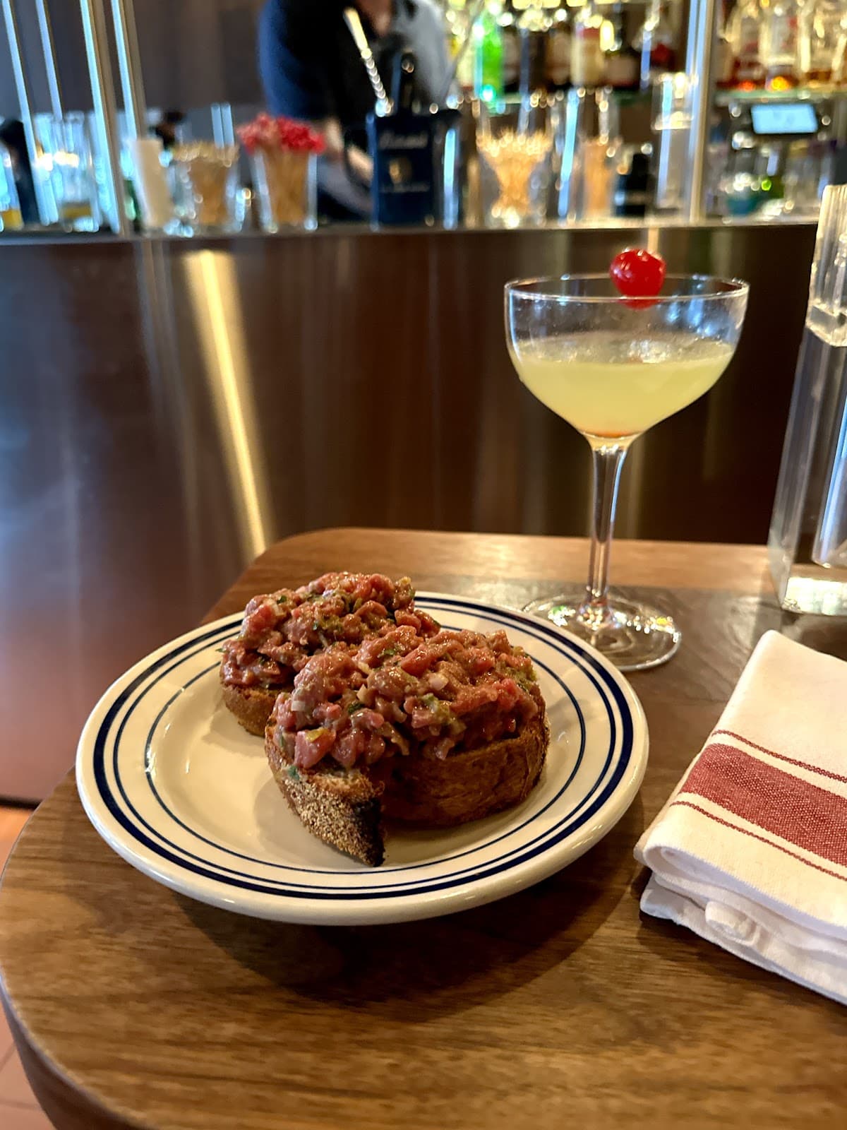 Close-up of cocktail and appetizer on bar counter with bartender in background in Toronto