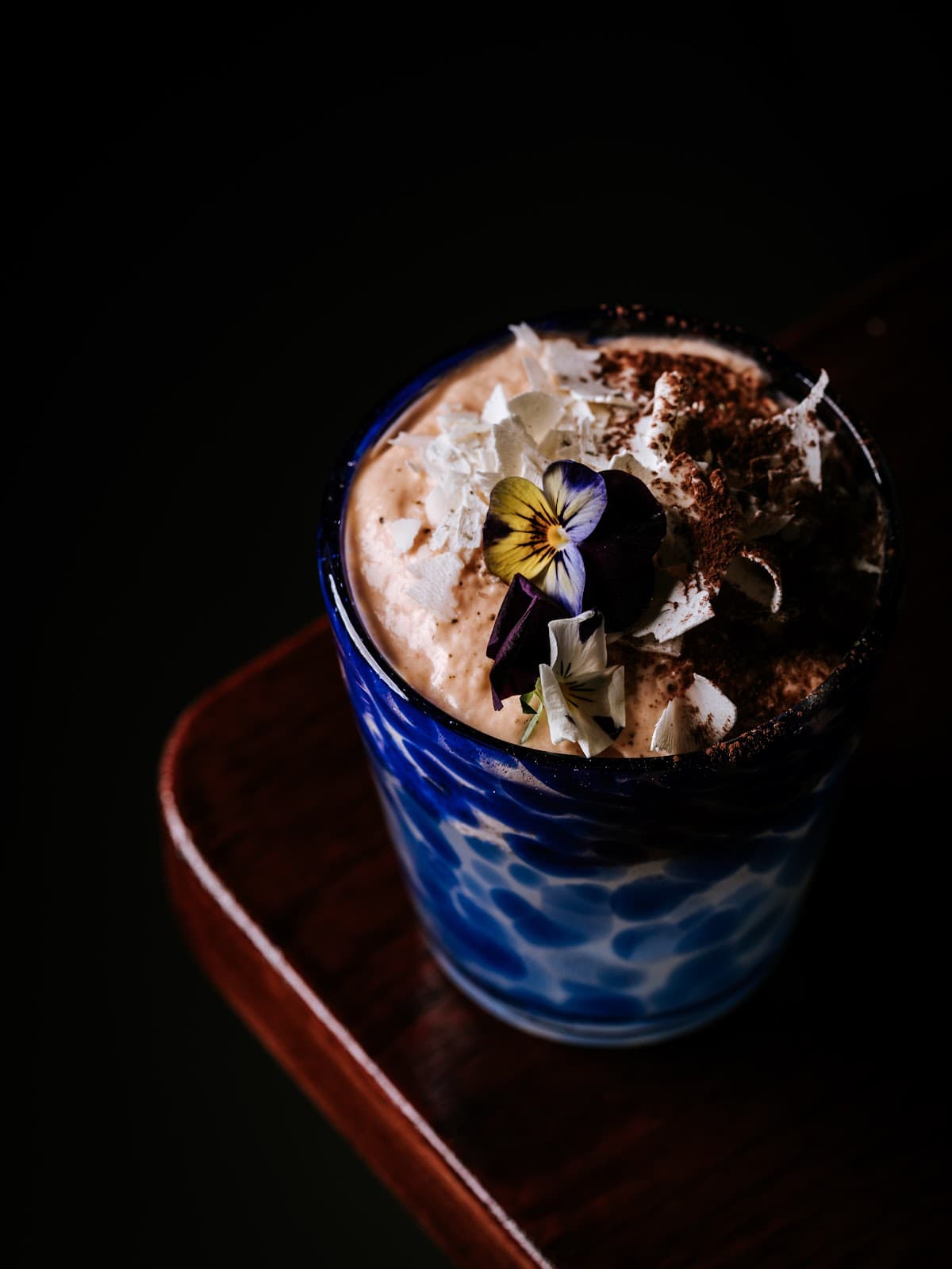 Artisanal cocktail with edible flowers in a blue-patterned glass on a dark wooden surface in Lisboa