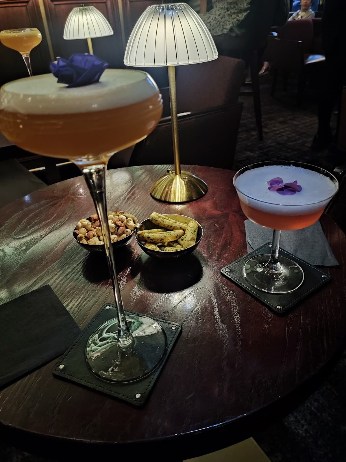 Two cocktails with foam and flowers on a wood table with snacks and lamp in London