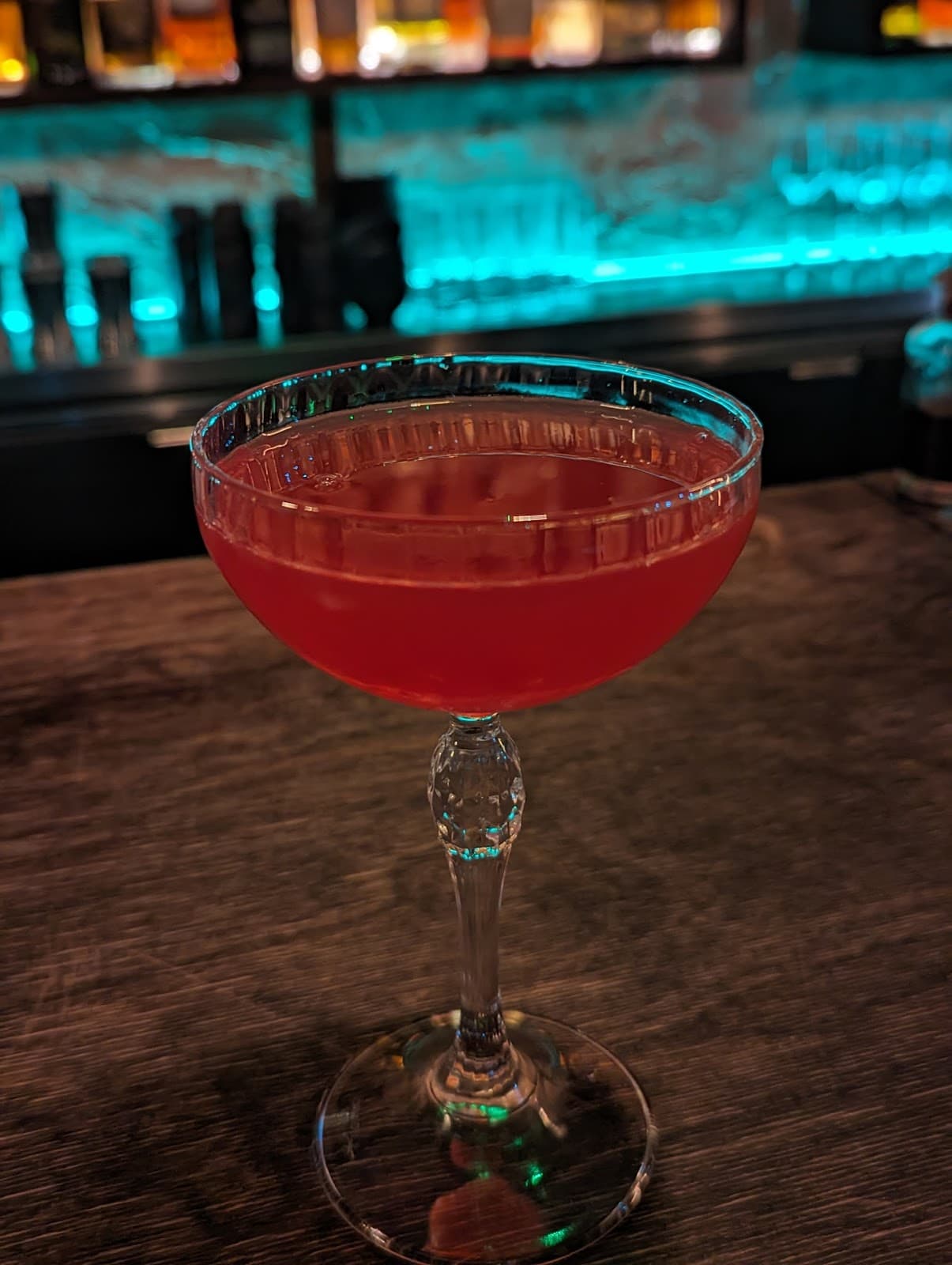 Red cocktail in a coupe glass on a bar counter with illuminated bottles in the background in London