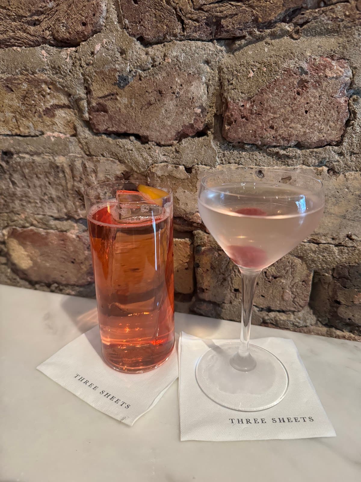 Two cocktails on a marble counter at Three Sheets with a brick wall background in London