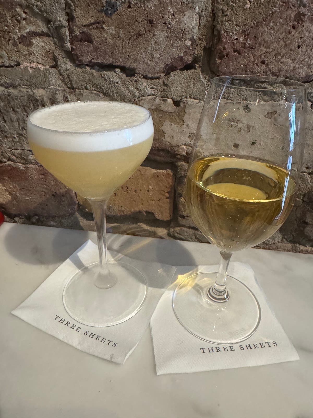 Two cocktails on a marble counter at Three Sheets with brick wall background in London