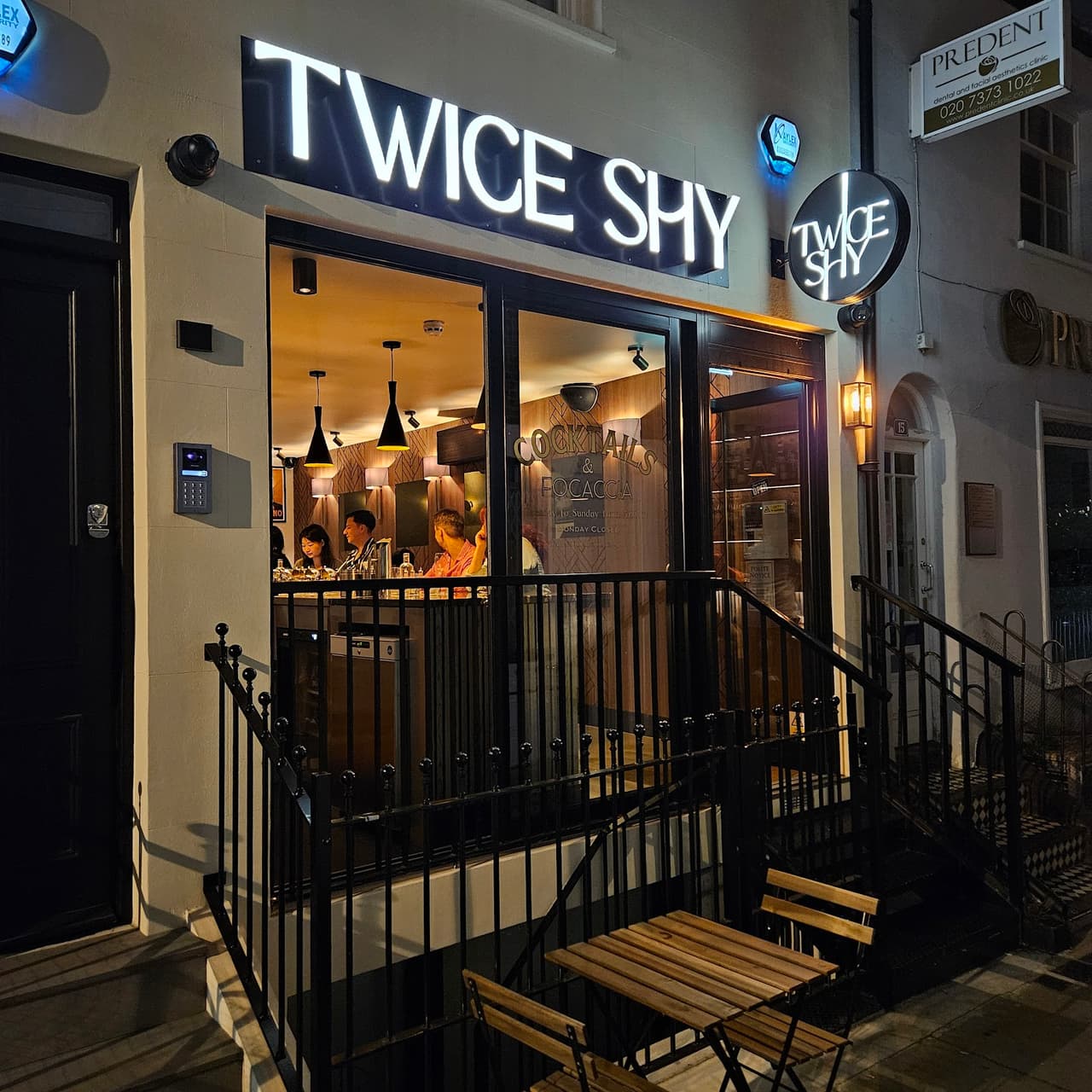 Exterior view of 'Twice Shy' cocktail bar with illuminated sign and outdoor seating